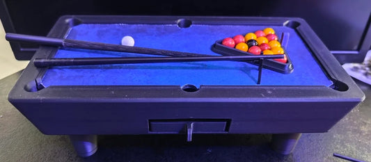 Billard miniature imprimé en trois dimensions, tapis bleu découpé dans un tapis de souris, deux queues sombres posées en travers de la table, bille blanche et triangle de billes de verre colorées