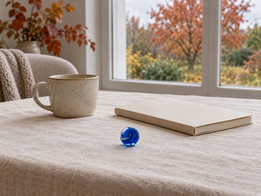 Une bille de verre bleue posée sur une nappe de lin écru, près d'un cahier fermé et d'une tasse en grès, devant une fenêtre ouverte sur un jardin aux arbres roux.