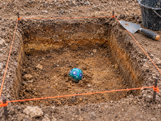 Une bille de verre turquoise à pastilles blanches et bleues, à demi dégagée au fond d'un carré de fouille bordé d'un cordeau orange, une truelle et un seau noir posés au bord.