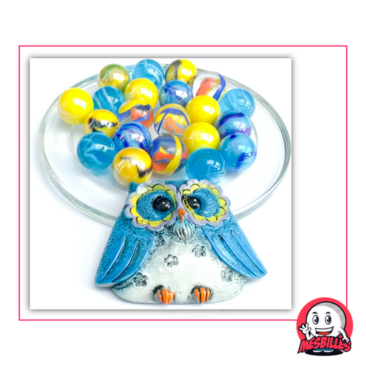 Blue ceramic owl accompanied by 20 Marbles of 16 mm in blue, Yellow, transparent and marbled glass, placed on a glass dish with pink frame and My-Glassmarbles Logo.